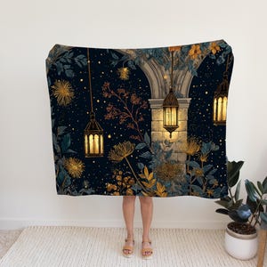 May include: A dark blue blanket with a whimsical illustration of a starry night scene. The design features glowing lanterns, floral elements, and an arched architectural structure. The blanket is draped over a person.