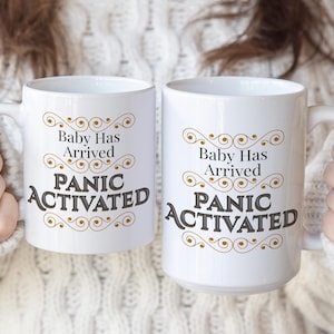 May include: Two white ceramic mugs featuring the text Baby Has Arrived and PANIC ACTIVATED in black lettering. The design includes decorative gold swirl flourishes above and below the text, centered on the front of each mug with a handle.