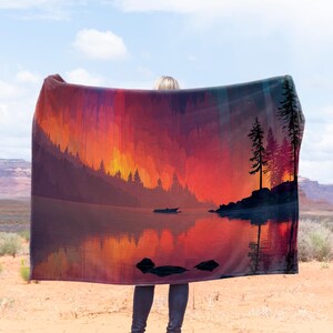 May include: A landscape blanket featuring a serene lake scene with a boat, silhouetted trees, and a vibrant sunset. The blanket's design includes a reflection of the trees and sky in the water, creating a tranquil and colorful image.