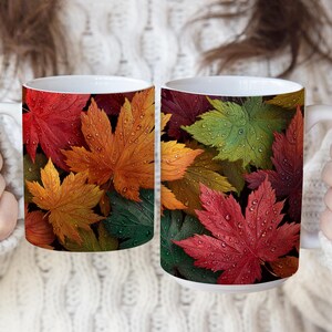 May include: Two white ceramic mugs with a vibrant autumn leaf design. The mugs feature colorful leaves in shades of red, orange, yellow, and green, with water droplets. The mugs are held by a person wearing a white sweater.