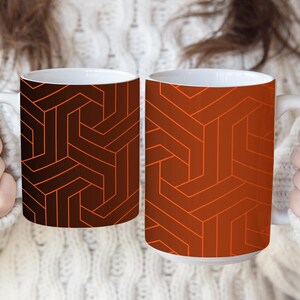 May include: Two ceramic mugs featuring a geometric pattern of interlocking lines. The left mug has a dark brown background with orange lines, while the right mug has a burnt orange background with darker orange lines. Both mugs have white interiors.