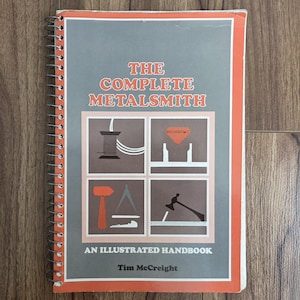 以下が含まれることがあります： 「The Complete Metalsmith」というタイトルの、オレンジとグレーの表紙のリング綴じの本。表紙には、金属加工のツールと技術のイラストが描かれています。著者の名前、Tim McCreightも表示されています。