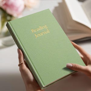 May include: A sage green Reading Journal with gold lettering. The hardcover book is held open, with the title visible. The journal is surrounded by a soft, neutral background, with a bouquet of pink flowers in the background.