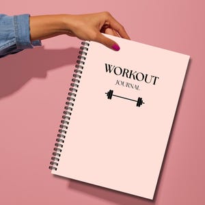 May include: A light pink spiral-bound workout journal with black text that reads WORKOUT JOURNAL and a black barbell icon centered on the front cover. The notebook features a black metal coil binding along the left side for page turning.