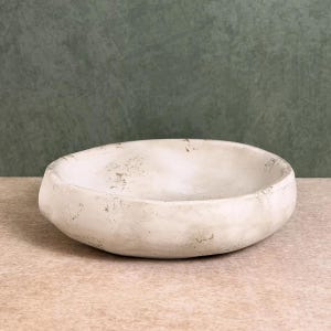 Pode incluir: Uma tigela circular rasa com uma superfície texturizada em tom esbranquiçado. O exterior e o interior apresentam um acabamento mate com subtis manchas castanhas de aspeto envelhecido e um rebordo irregular e orgânico de aspeto artesanal.