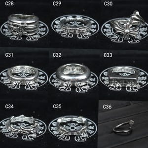 Puede incluir: Una colecci&oacute;n de nueve anillos de plata etiquetados del C28 al C36, con varios dise&ntilde;os intrincados que incluyen motivos de cruz, bandas grabadas y estilos abiertos. Cada anillo se muestra individualmente sobre una superficie negra con logo.