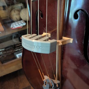 May include: A close-up of a dark brown upright bass with strings and a gray string holder. The string holder has the text "NEAR ART CO." printed on it. The bass has a wooden bridge and f-holes.