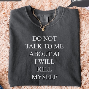 May include: Pepper-colored t-shirt with the text "DO NOT TALK TO ME ABOUT AI I WILL KILL MYSELF" in white. A gold chain necklace rests on the shirt. The shirt is folded on a fluffy, peach-colored surface.