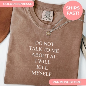 May include: An espresso-colored short-sleeve t-shirt with a crew neckline. The shirt features white text printed on the front that reads: DO NOT TALK TO ME ABOUT AI I WILL KILL MYSELF. A silver-toned necklace with a small round pendant rests on top.