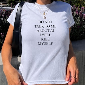 May include: A white short-sleeved t-shirt featuring black text printed on the front that reads: DO NOT TALK TO ME ABOUT AI I WILL KILL MYSELF. The shirt has a crew neckline and is worn with a layered gold-tone necklace and a black shoulder bag.