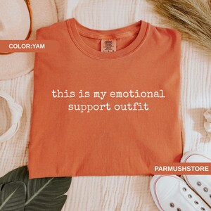 May include: A yam-colored t-shirt with the text "this is my emotional support outfit" in white. The shirt is made of a soft material and is displayed on a white surface. White sneakers are visible in the bottom right corner.