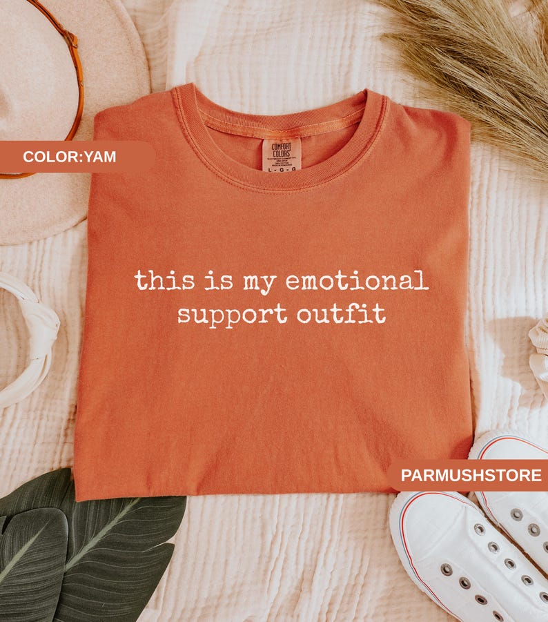 May include: A yam-colored t-shirt with the text "this is my emotional support outfit" in white. The shirt is made of a soft material and is displayed on a white surface. White sneakers are visible in the bottom right corner.