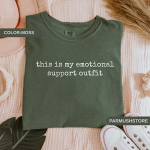 May include: Moss green t-shirt with the text "this is my emotional support outfit" in white. The shirt is folded on a light pink surface, with white sneakers and decorative elements in the background.