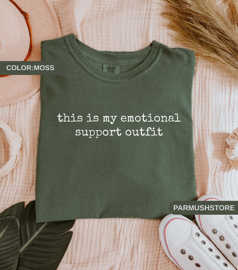 May include: Moss green t-shirt with the text "this is my emotional support outfit" in white. The shirt is folded on a light pink surface, with white sneakers and decorative elements in the background.