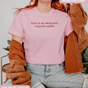 May include: A light pink t-shirt with the text "this is my emotional support outfit" in a dark red font. The shirt is paired with light blue jeans and a brown jacket.