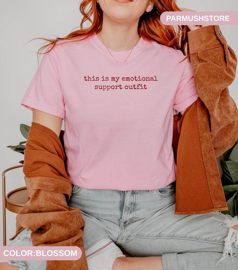 May include: A light pink t-shirt with the text "this is my emotional support outfit" in a dark red font. The shirt is paired with light blue jeans and a brown jacket.