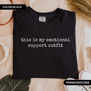 May include: Black crew neck sweatshirt with the text "this is my emotional support outfit" in white. The sweatshirt is displayed on a beige surface with a pair of white sneakers and a hat.