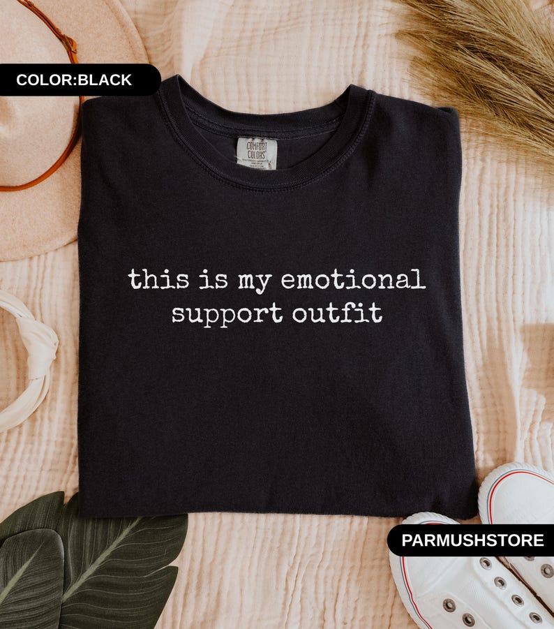 May include: Black crew neck sweatshirt with the text "this is my emotional support outfit" in white. The sweatshirt is displayed on a beige surface with a pair of white sneakers and a hat.