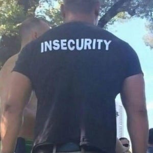 May include: A black t-shirt with the word "INSECURITY" printed in white block letters across the back. The shirt is worn by a person, with the focus on the text and the back of the shirt.
