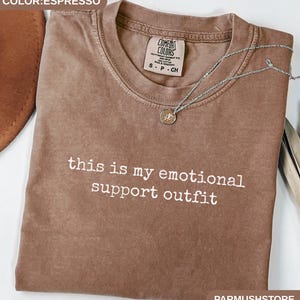 May include: Espresso-colored t-shirt with the text "this is my emotional support outfit" in white. A silver necklace with a small pendant rests on the shirt. The shirt is from Comfort Colors.