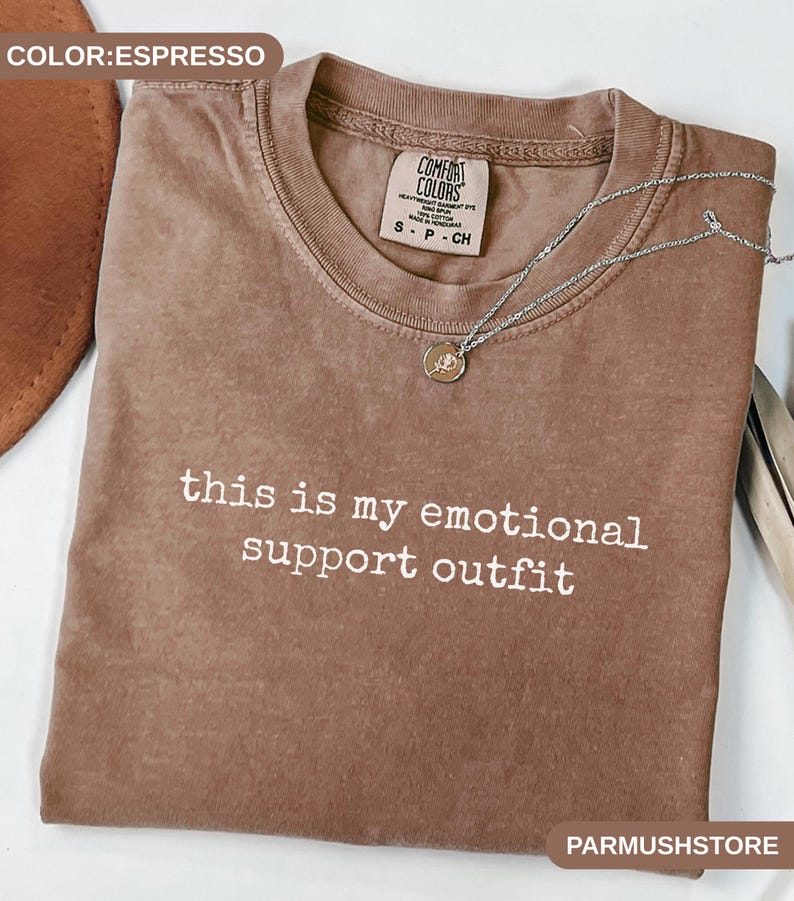 May include: Espresso-colored t-shirt with the text "this is my emotional support outfit" in white. A silver necklace with a small pendant rests on the shirt. The shirt is from Comfort Colors.