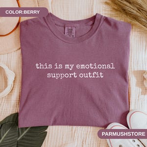 May include: Berry-colored t-shirt with the text "this is my emotional support outfit" in white. The shirt is a crew neck style. The image also shows a pair of white sneakers.
