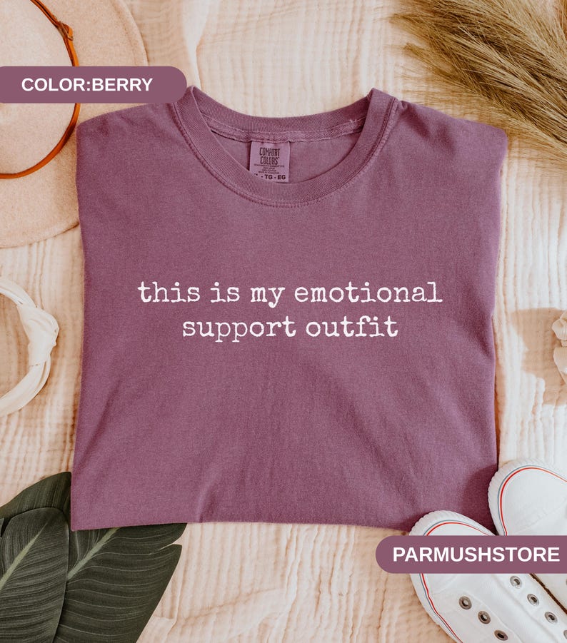 May include: Berry-colored t-shirt with the text "this is my emotional support outfit" in white. The shirt is a crew neck style. The image also shows a pair of white sneakers.