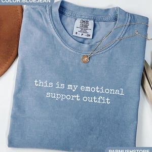 May include: Bluejean-colored t-shirt with the text "this is my emotional support outfit" in white. A gold necklace with a pendant rests on the shirt. The shirt is folded and has a Comfort Colors tag.
