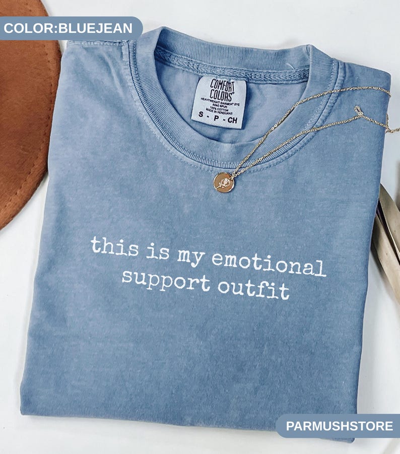 May include: Bluejean-colored t-shirt with the text "this is my emotional support outfit" in white. A gold necklace with a pendant rests on the shirt. The shirt is folded and has a Comfort Colors tag.