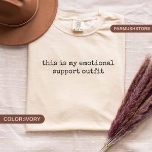 May include: Ivory-colored t-shirt with the text "this is my emotional support outfit" in black. The shirt is a Comfort Colors XL size. A brown hat and dried flowers are also in the image.