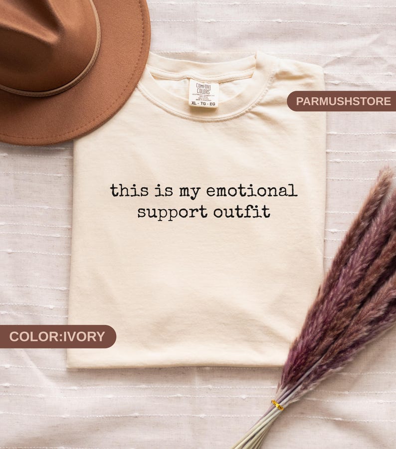 May include: Ivory-colored t-shirt with the text "this is my emotional support outfit" in black. The shirt is a Comfort Colors XL size. A brown hat and dried flowers are also in the image.