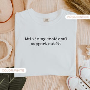 May include: White t-shirt with the text "this is my emotional support outfit" in black. The shirt is laid flat on a beige surface, with white sneakers visible at the bottom. The shirt has a Comfort Colors tag.