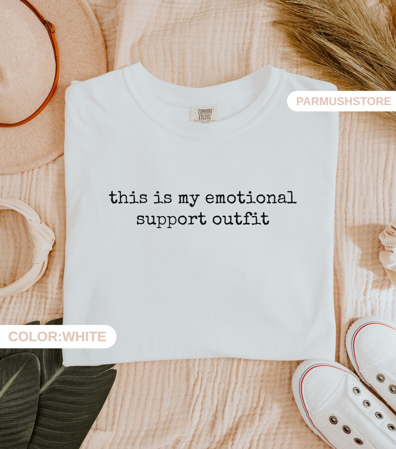 May include: White t-shirt with the text "this is my emotional support outfit" in black. The shirt is laid flat on a beige surface, with white sneakers visible at the bottom. The shirt has a Comfort Colors tag.