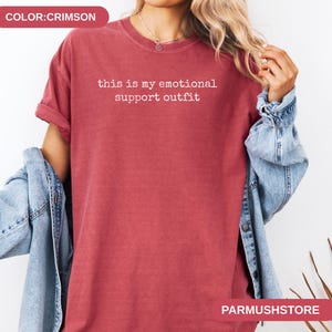 May include: Crimson-colored t-shirt with the text "this is my emotional support outfit" in white. The shirt has a crew neck and short sleeves. A denim jacket is draped over the shoulders.