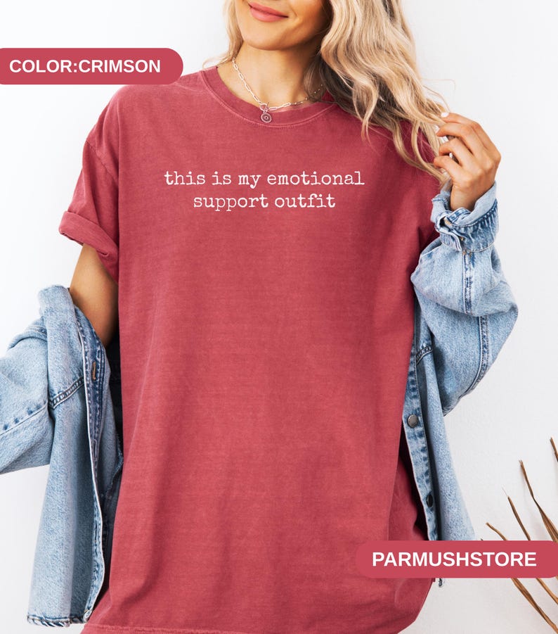 May include: Crimson-colored t-shirt with the text "this is my emotional support outfit" in white. The shirt has a crew neck and short sleeves. A denim jacket is draped over the shoulders.