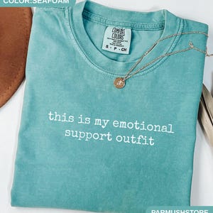 May include: Seafoam green t-shirt with the text "this is my emotional support outfit" in white. A gold necklace with a pendant rests on the shirt. The shirt has a Comfort Colors tag.