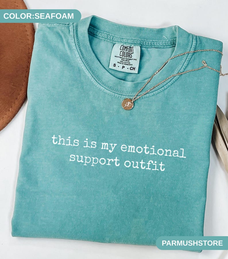 May include: Seafoam green t-shirt with the text "this is my emotional support outfit" in white. A gold necklace with a pendant rests on the shirt. The shirt has a Comfort Colors tag.