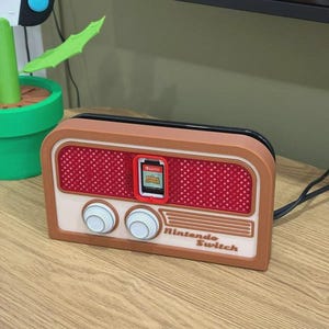 Op de afbeelding: Een bruine en crèmekleurige Nintendo Switch game cartridge houder in de vorm van een vintage radio. De houder heeft een rood luidsprekerrooster, twee witte knoppen en de tekst "Nintendo Switch". Een game cartridge is geplaatst.