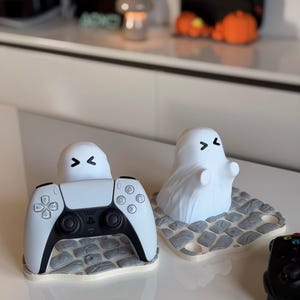 Puede incluir: Figuras blancas con forma de fantasma con rasgos faciales negros y una base gris con forma de piedra. Una figura está encima de un mando de juego blanco y negro. La otra es una figura de fantasma independiente. Tema: Halloween y juegos.