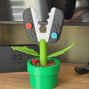 Può includere: Un vaso di plastica verde con interno marrone contiene uno stelo verde con due controller Switch attaccati, che assomigliano a una pianta. I controller sono grigi e bianchi con pulsanti rossi e blu. Due cartucce di gioco sono nel vaso.
