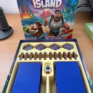 Può includere: Una scatola di gioco da tavolo intitolata "Survive the Island" con un'illustrazione colorata di persone su un'isola. La scatola del gioco è aperta, rivelando i pezzi del gioco, tra cui carte blu e pezzi di legno marrone.