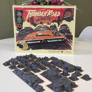 May include: A board game box titled "Thunder Road Vendetta" with a car graphic. Gray terrain pieces are arranged in front of the box. The box has a red and orange color scheme. The text "This road is paved with bad intentions!" is visible.