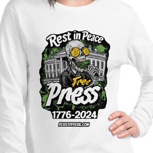 May include: White long-sleeve shirt featuring a graphic with the text "Rest in Peace Free Press 1776-2024". The graphic includes a skeleton wearing a suit and sunglasses, with the White House in the background. The website address is also visible.