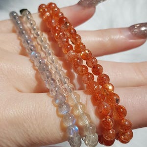 May include: Three beaded bracelets in shades of orange, clear, and grey. The bracelets are made of small, round beads. The orange bracelet has a sparkling appearance. The bracelets are displayed on a light background.