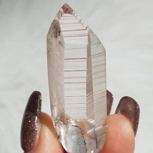 May include: A clear quartz crystal with internal lines and a pointed end. The crystal has a smoky look with some inclusions. It is held against a soft, blurred background. The crystal is a natural, unpolished gemstone, often used for its properties.