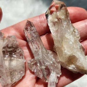 May include: Three clear quartz crystal points with natural facets and internal inclusions. The crystals are translucent with varying geometric shapes, sharp terminations, and a glassy texture, displaying natural mineral formations.