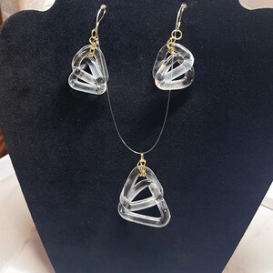 May include: A clear, geometric jewelry set featuring a necklace and matching earrings. The earrings have gold-colored hooks, and the necklace has a gold-colored pendant. The pendant and earrings are made of clear, interlocking triangular shapes.