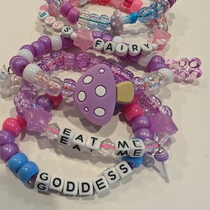 May include: A collection of colorful beaded bracelets in shades of purple, pink, and blue. Some bracelets feature letter beads spelling out words like "FAIRY", "EAT ME", and "GODDESS". One bracelet has a purple mushroom charm.