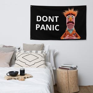 Puede incluir: Tapiz de pared negro con el texto blanco "DONT PANIC". El tapiz presenta un personaje de dibujos animados con cabello naranja y una chaqueta naranja. El personaje tiene una expresión preocupada.