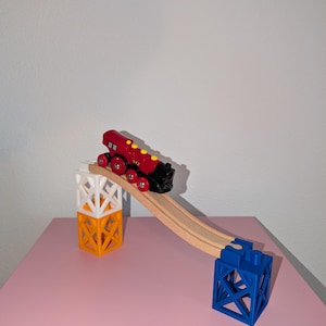 Könnte beinhalten: Ein rot-schwarzer Spielzeugzug mit gelben Akzenten fährt eine Holzstrecke hinunter, die von weißen, orangefarbenen und blauen geometrischen Strukturen getragen wird. Das Spielzeugzug-Set befindet sich auf einer rosa Oberfläche.